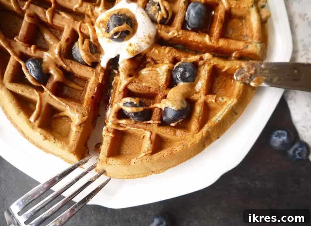 A stack of Paleo gluten-free mashed blueberry waffles on a white plate, with a side of fresh blueberries and maple syrup.