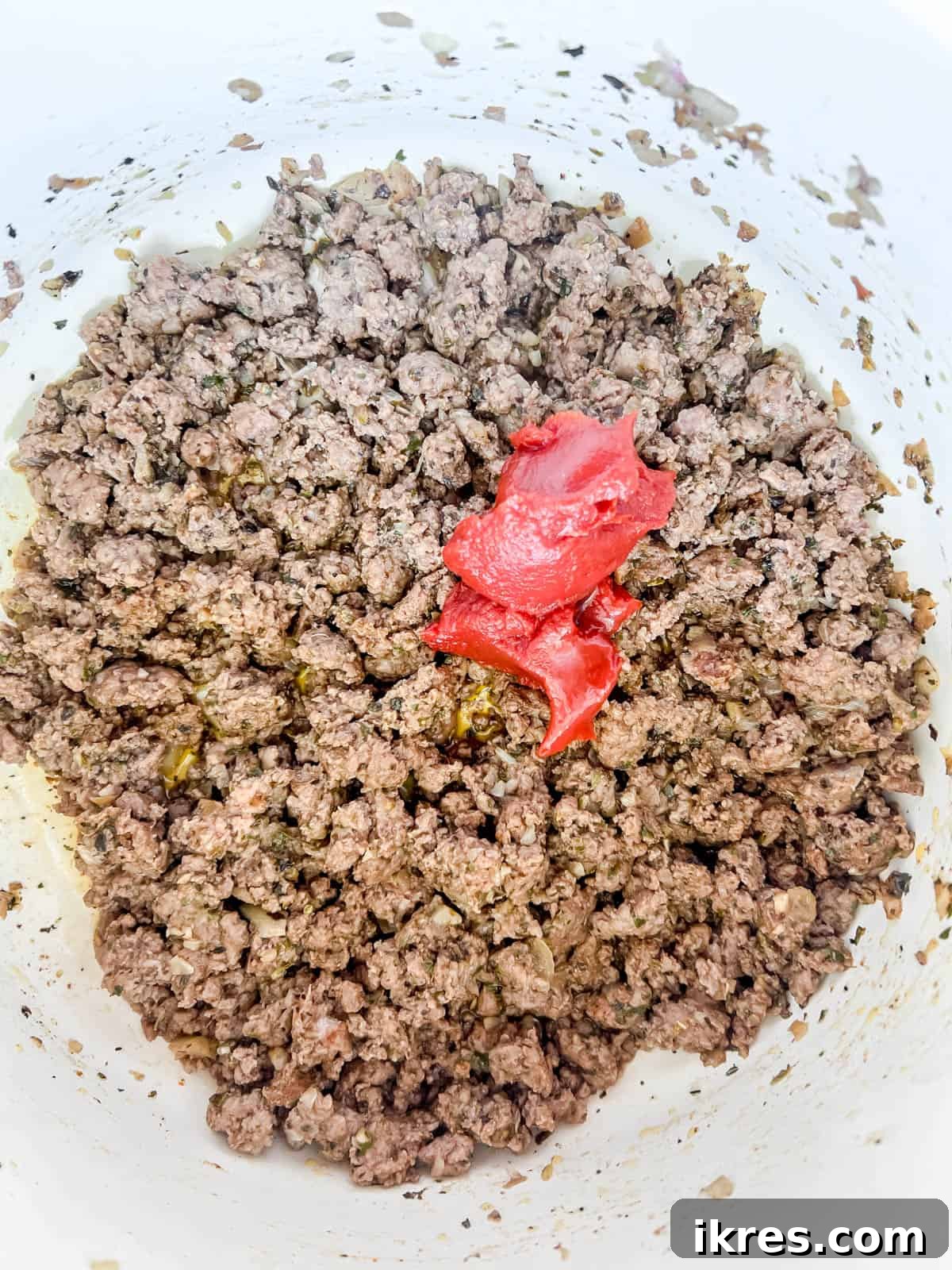 Tomato paste being stirred into the browned ground beef mixture in a pan.