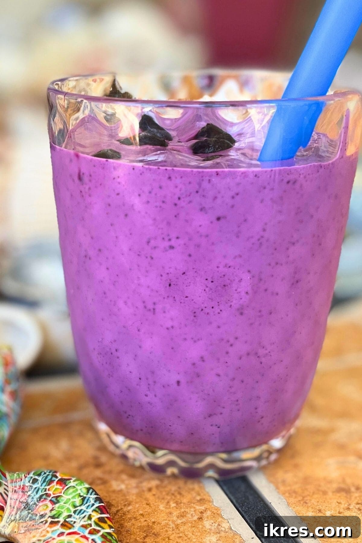 A bluberry banana and peanut butter smoothie in a clear glass on a tiled background with a blue straw and shells behind it.