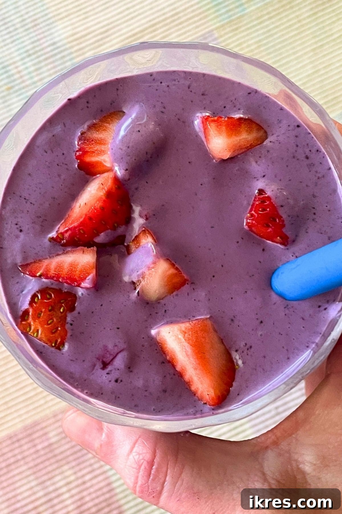 A blueberry banana and peanut butter smoothie with a blue straw held in a hand in a clear galss topped with some diced strawberries.