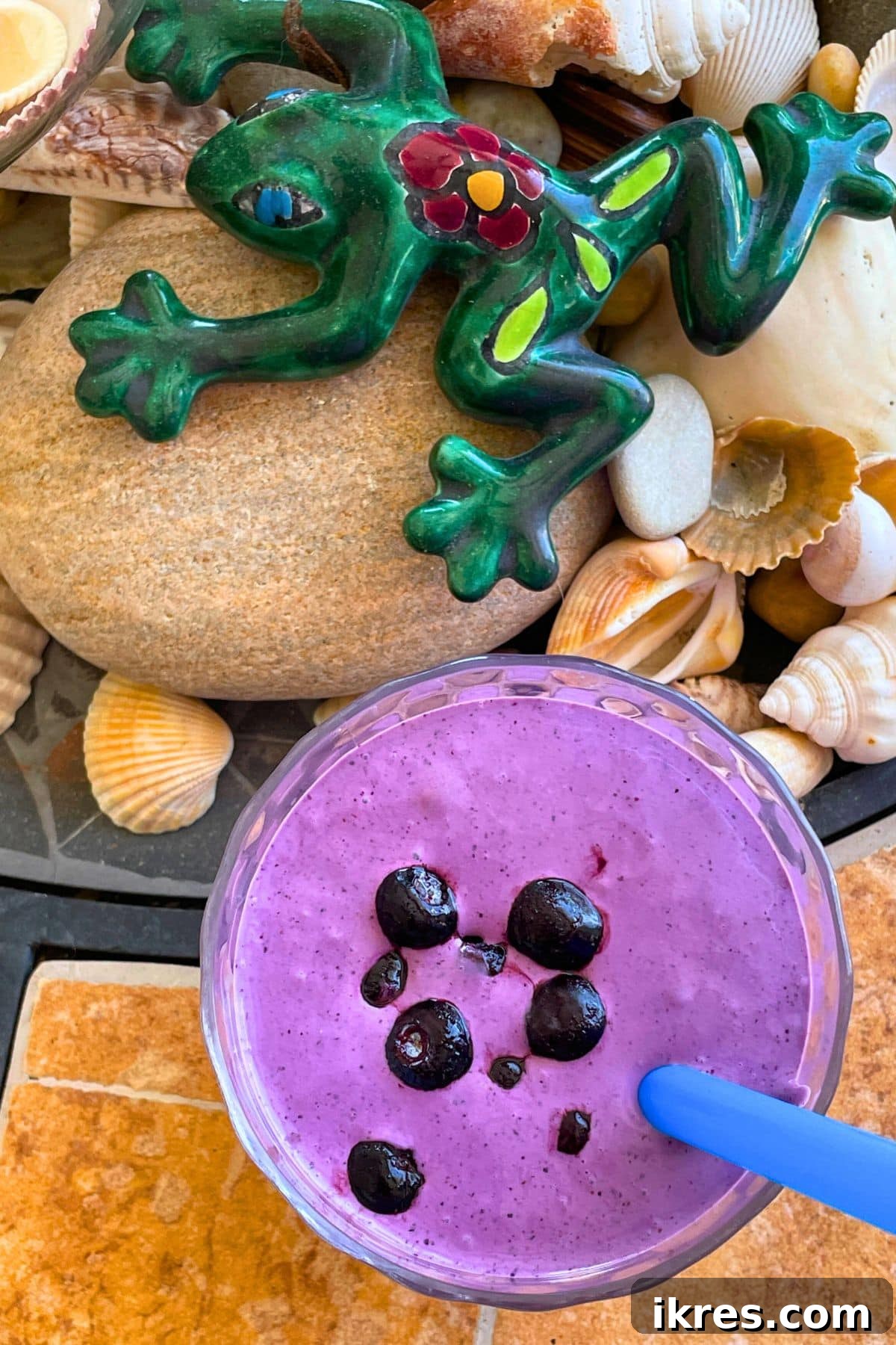 A bluberry banana and peanut butter smoothie with blueberries on top on a tiled background with a blue straw and a patterned frog and shells off to the side.