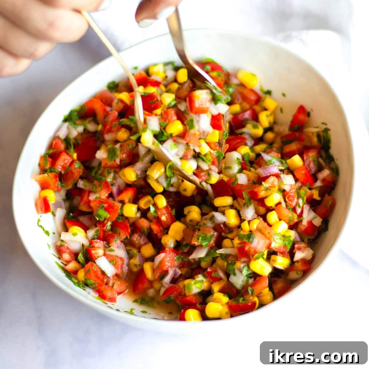Zesty Sweet Corn Pico de Gallo 5 Two golden serving spoons actively tossing a freshly prepared bowl of corn pico de gallo, mixing the vibrant ingredients together.