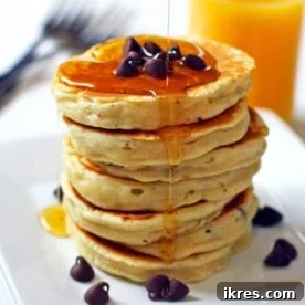 A stack of chocolate chip pancakes with maple syrup on a white plate with a glass of orange juice in the background.