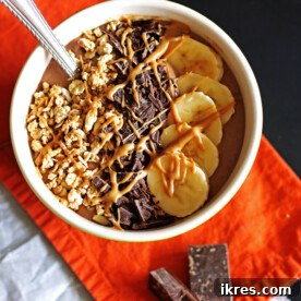 A vibrant chocolate peanut butter smoothie bowl topped with sliced bananas, nuts, and chocolate shavings.