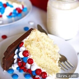A delectable slice of M&M Kit Kat Cake on a white dessert plate, with a clear glass of milk nearby.