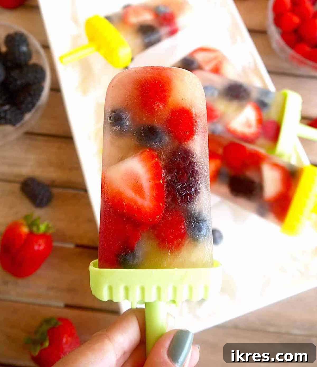 Two vibrant apple juice popsicles, studded with colorful berries, held in hands against a sunny background.