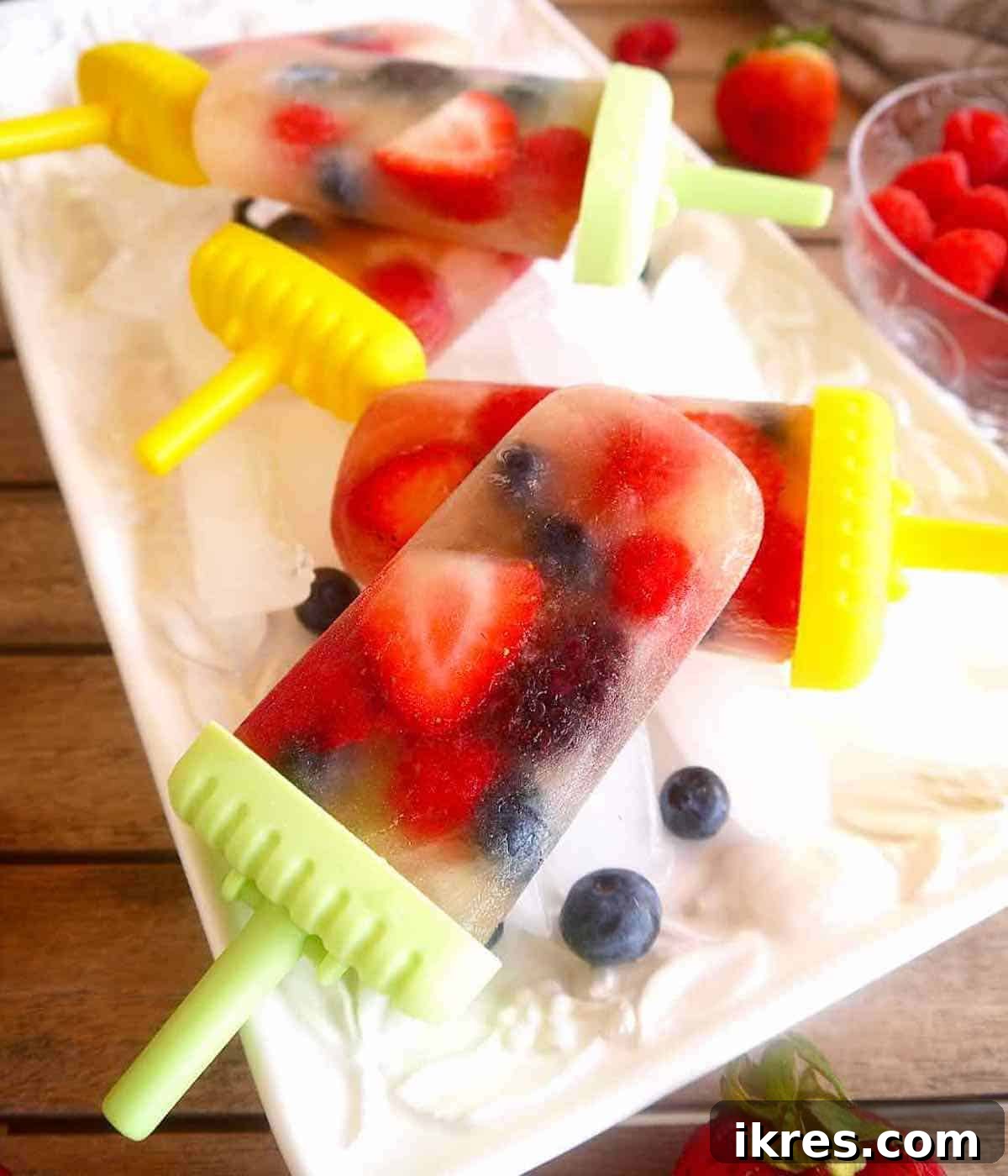 A selection of colorful apple juice popsicles with mixed berries arranged on a bed of ice.
