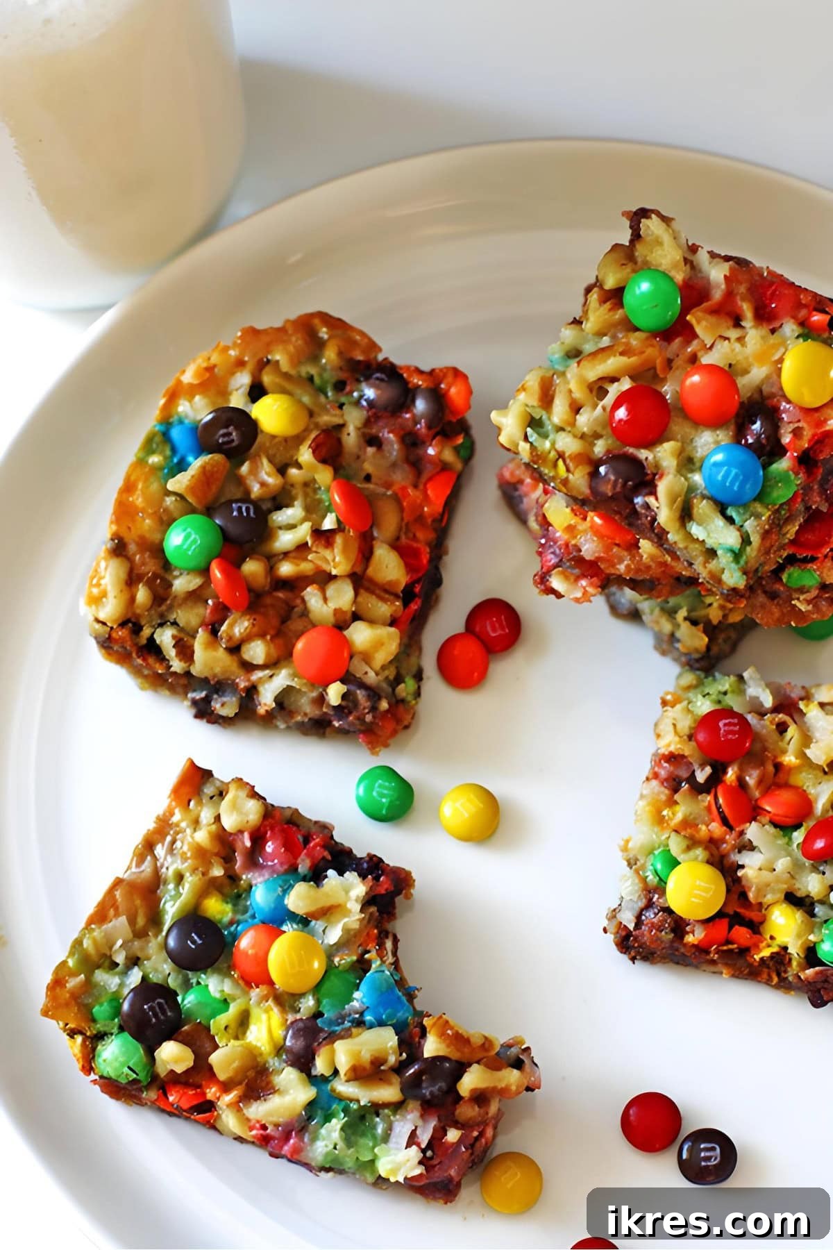 Wondrous Seven-Layer Magic Bars 2 Four magic cookie bars with walnuts, coconut, chocolate, and mini m and ms on a white plate with a glass of milk.