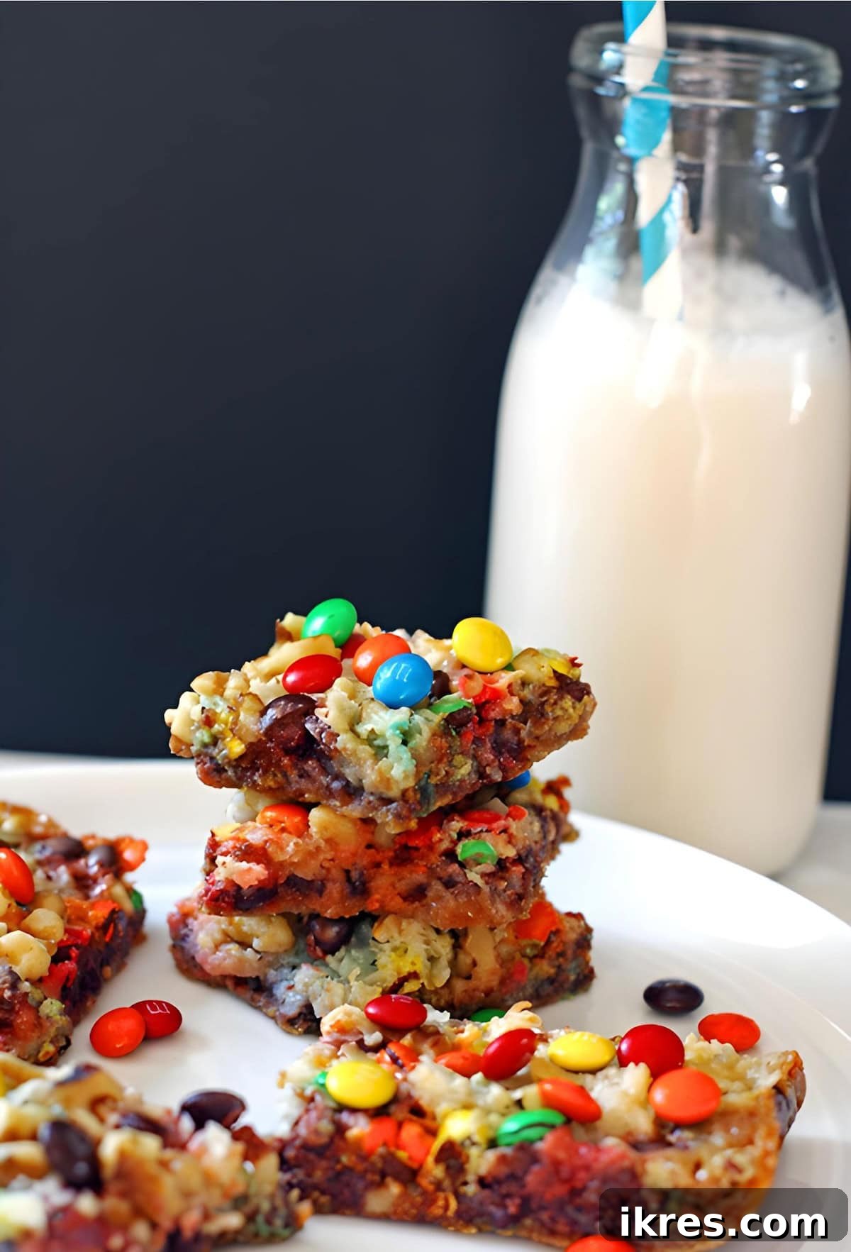 Wondrous Seven-Layer Magic Bars 5 Four 7-layer cookie bars with nuts, coconut, chocolate chips, and mini m and ms stacked on a white plate with a glass of milk.