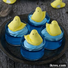 Close-up of a clear cup filled with blue Jello, topped with whipped cream and a yellow marshmallow Peep chick, designed as a 'Swimming Peeps Jello Cup'.