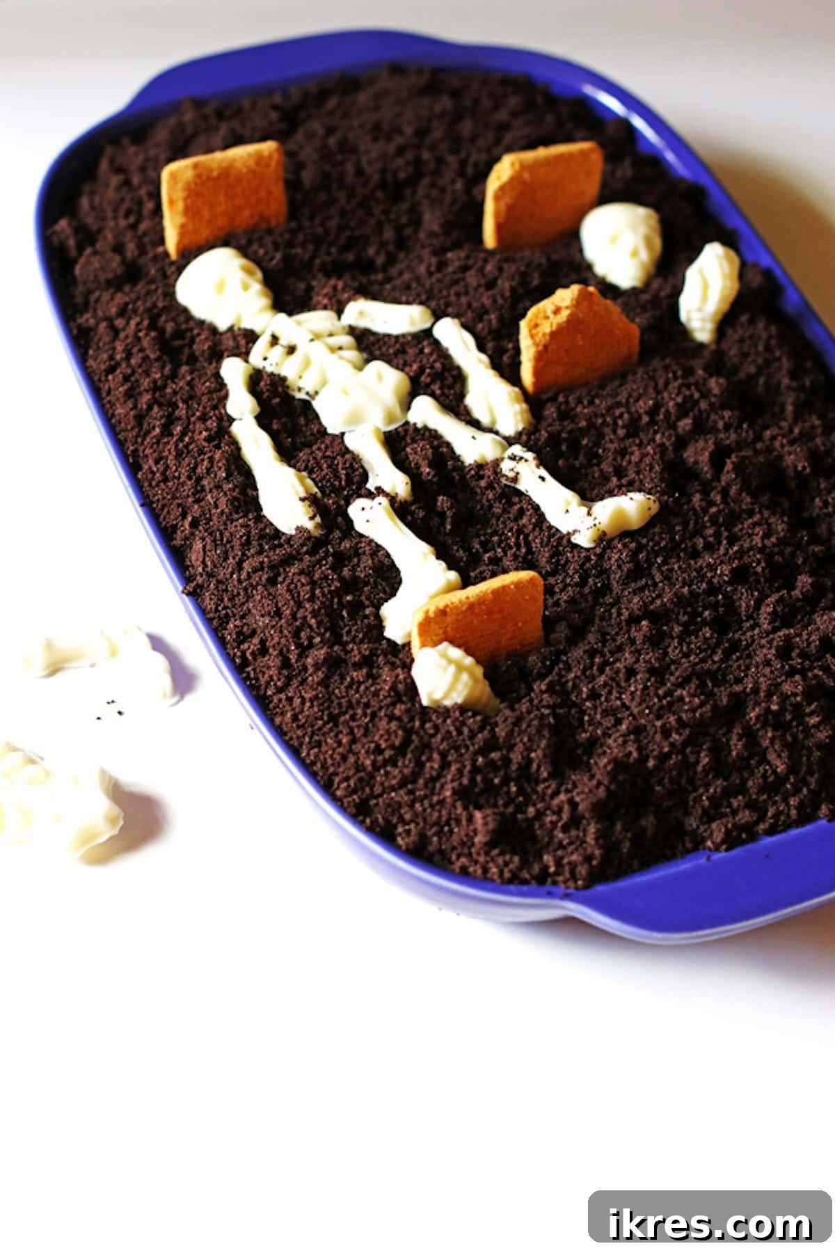 Festive Dirt Cake for Halloween and Easter 8 The Halloween Dirt Cake being adorned with white chocolate skeleton pieces and rectangular cookies artfully crafted to resemble tombstones, enhancing its spooky graveyard theme.