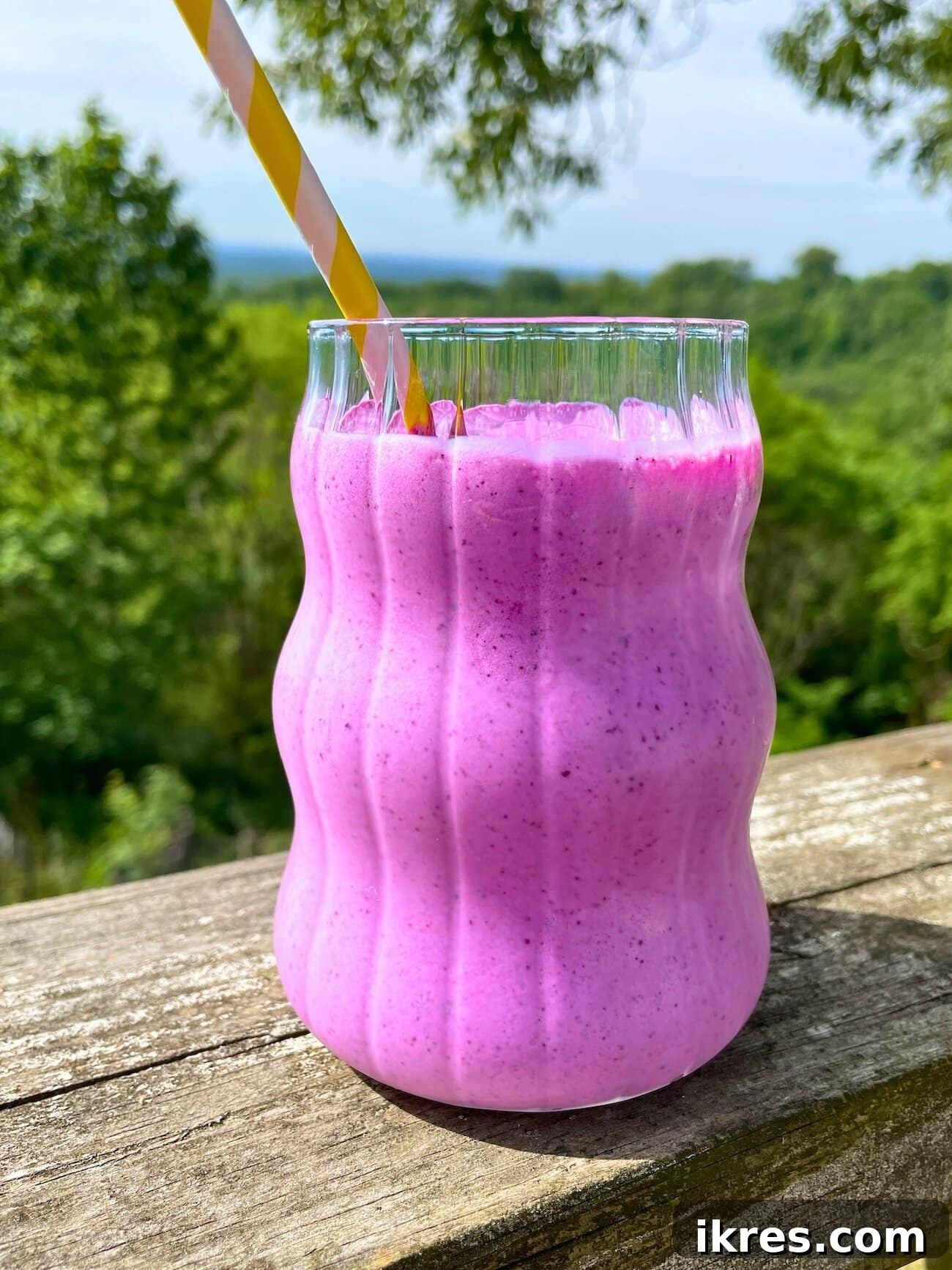 Vibrant Blueberry Pineapple Blast 2 A vibrant blueberry pineapple smoothie in a clear glass with a yellow straw, resting on a wooden railing outdoors with lush green trees in the background. The scene suggests a refreshing, healthy drink enjoyed in nature.