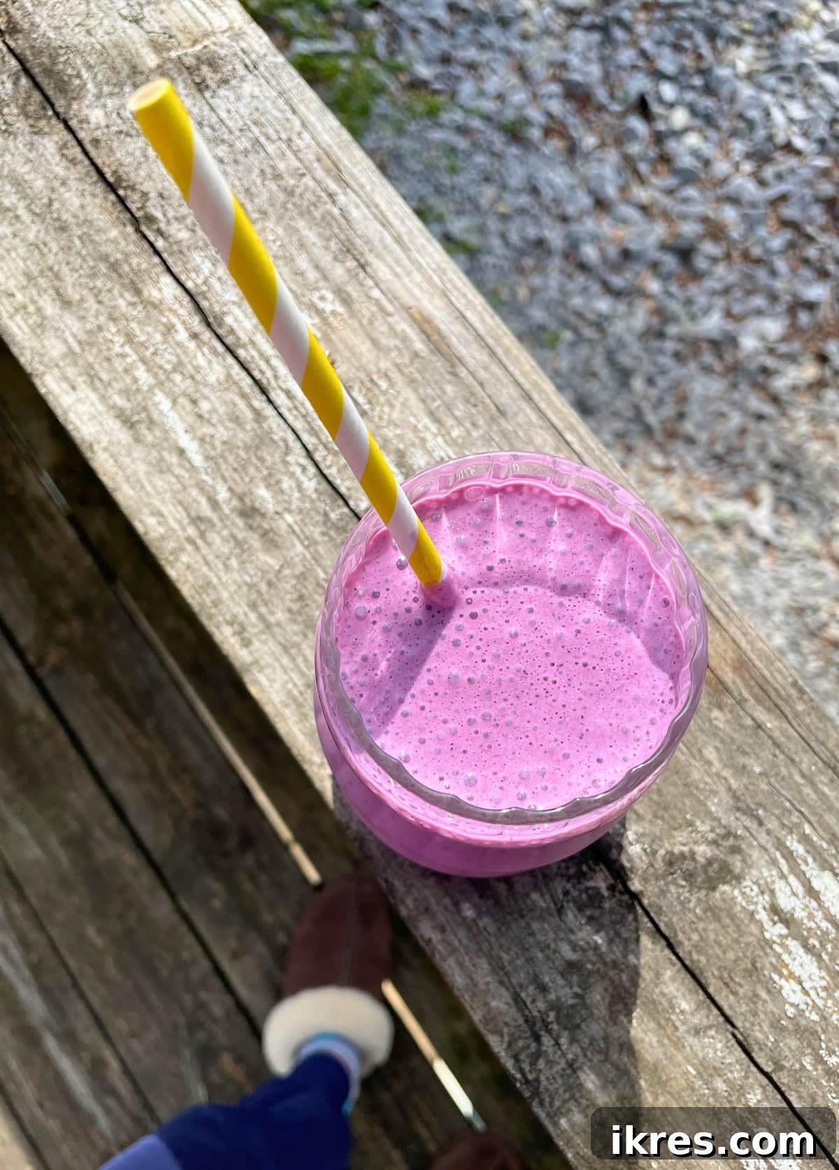 Vibrant Blueberry Pineapple Blast 9 A beautifully presented blueberry pineapple smoothie with a white and yellow striped straw, sitting on a wooden railing overlooking a scenic gravel driveway, perfect for a peaceful breakfast.