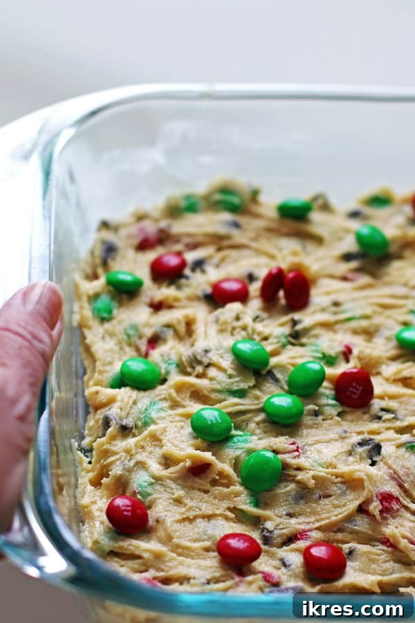 Decadent Cookie Squares 4 Adding red and green M&M candies to the top of the cookie bar batter in a glass baking pan before baking.