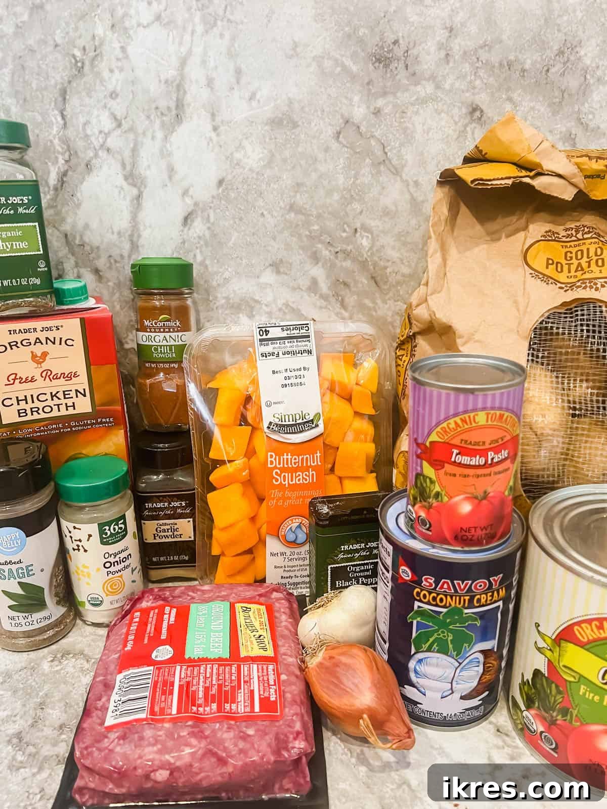 A flat lay photograph showcasing all the fresh and whole ingredients neatly arranged for preparing butternut squash shepherd's pie, including a whole squash, golden potatoes, ground meats, various herbs, and other vegetables.