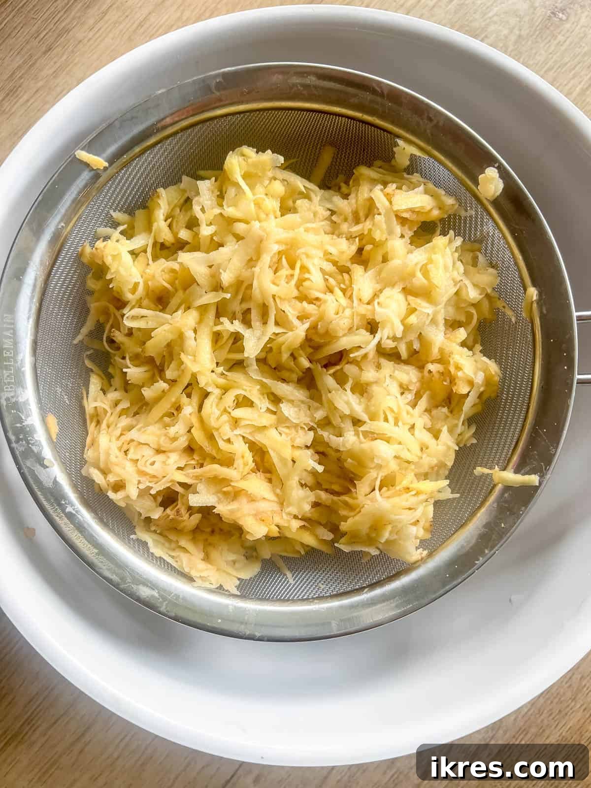 Raw shredded potatoes are placed in a sieve over a bowl to drain excess liquid after being mixed with salt.
