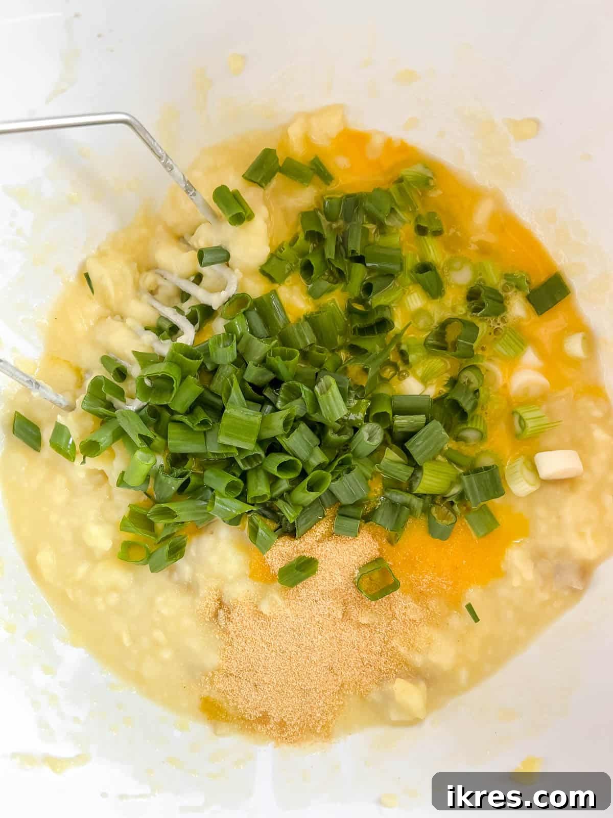 Minced garlic, beaten eggs, and sliced green onions are being added to a bowl of mashed potatoes.