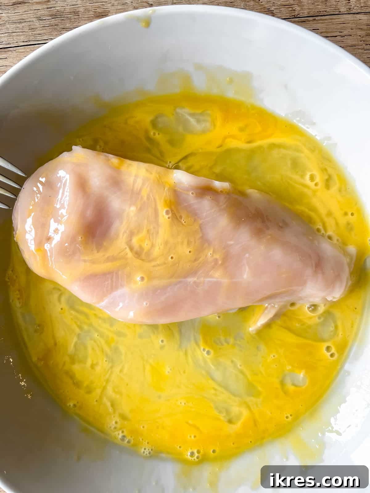 A boneless, skinless chicken breast being fully submerged and coated in the whisked egg wash.