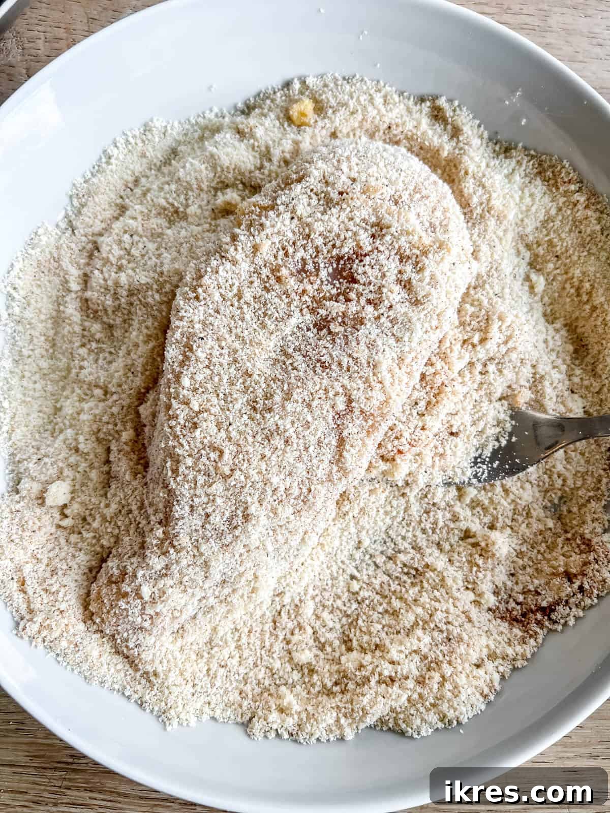 An egg-coated chicken breast being pressed firmly into the almond flour and spice mixture to achieve a complete breading.
