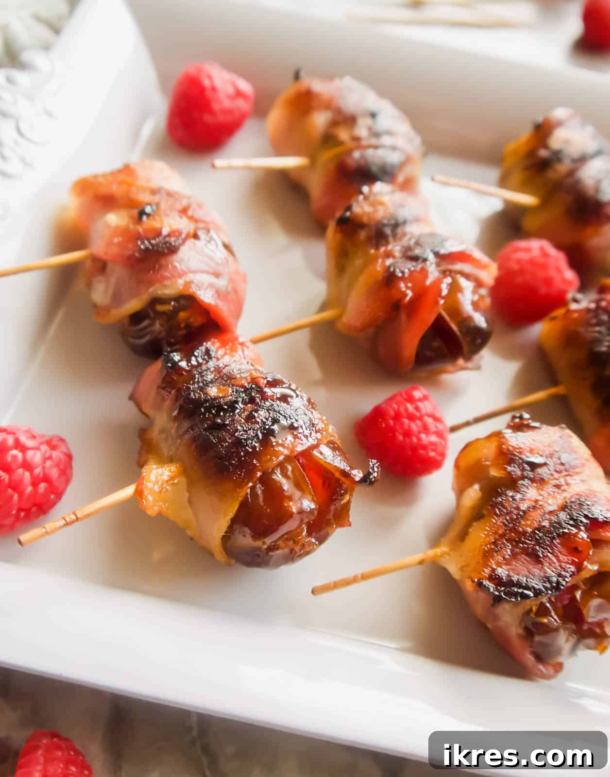 Blissful Bacon Wrapped Raspberry Date Bites 2 A plate featuring golden-brown, crispy bacon-wrapped dates, each filled with a burst of raspberry. Fresh raspberries are scattered around the plate, enhancing the visual appeal.