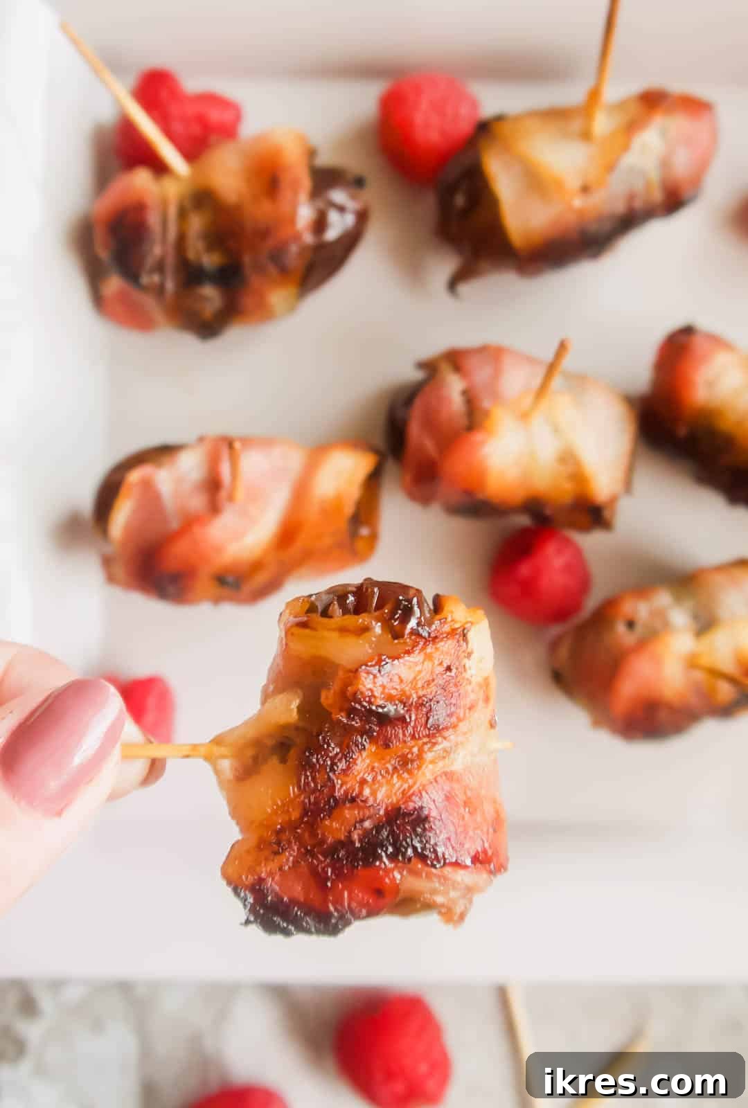 Blissful Bacon Wrapped Raspberry Date Bites 3 A single Raspberry Stuffed Medjool Date Wrapped in Crispy Bacon, being lifted from a plate by a toothpick, showcasing its perfect golden-brown exterior.