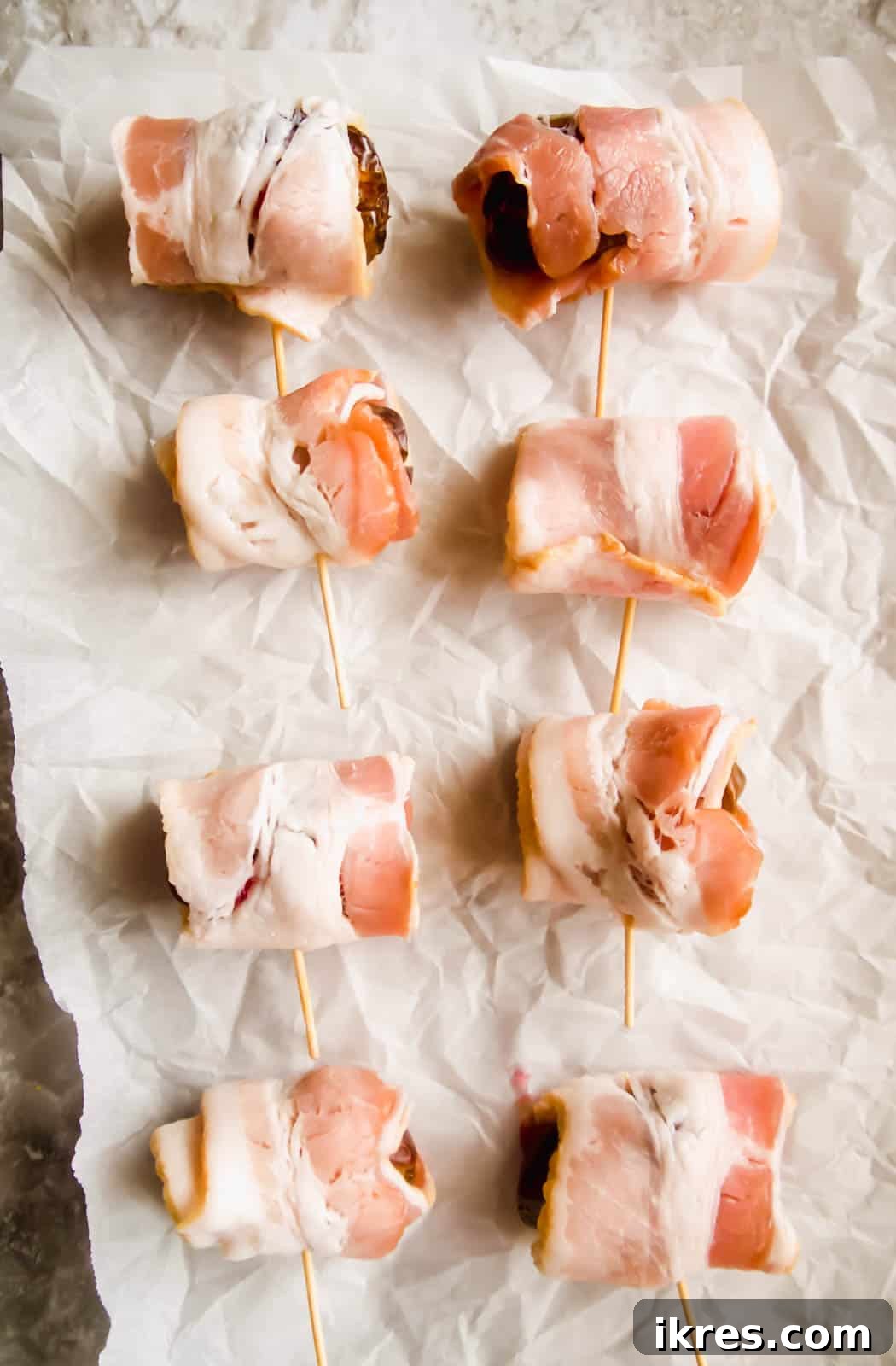 Blissful Bacon Wrapped Raspberry Date Bites 7 Raspberry-stuffed dates, each meticulously wrapped with a strip of raw bacon and secured with a toothpick, laid out on a baking sheet before cooking.