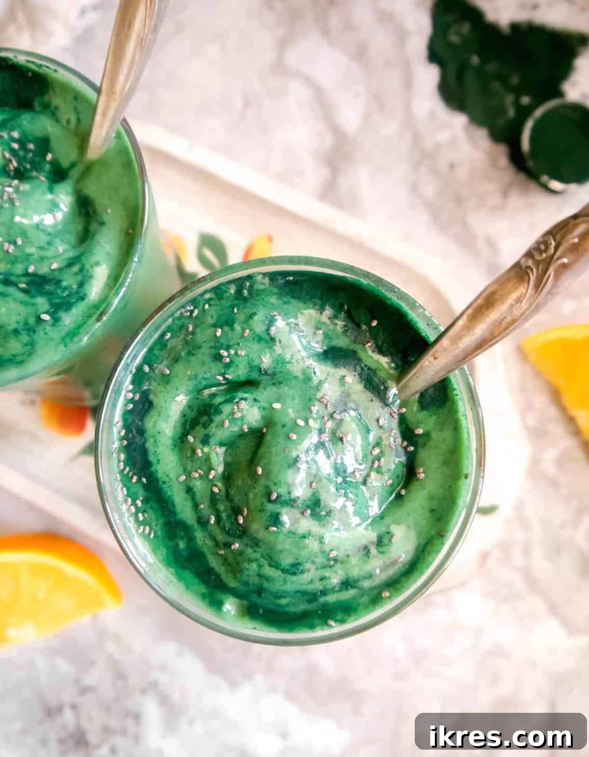 Sunrise Spirulina Fusion 2 Two glasses of a vibrant green spirulina smoothie, garnished with chia seeds. Fresh oranges and mango are visible in the background, hinting at the smoothie's ingredients.