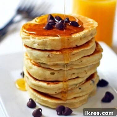 Golden Choc-Chip Stacks 5 A stack of chocolate chip pancakes with maple syrup on a white plate with a glass of orange juice in the background.
