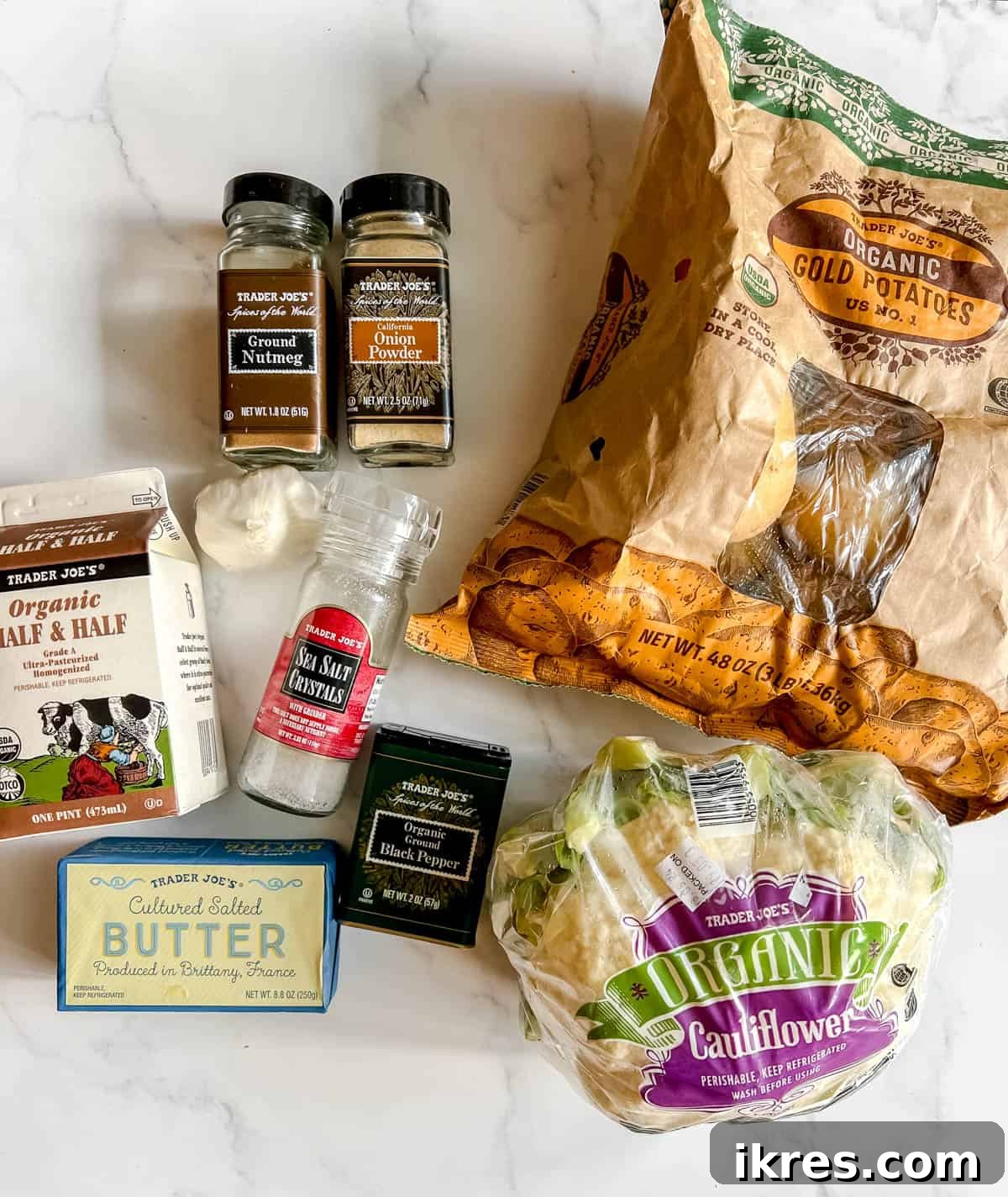 A flat lay photograph showcasing all the fresh ingredients needed to make the cauliflower and potato mash, including whole cauliflower, gold potatoes, garlic cloves, butter, and a carton of half-and-half.