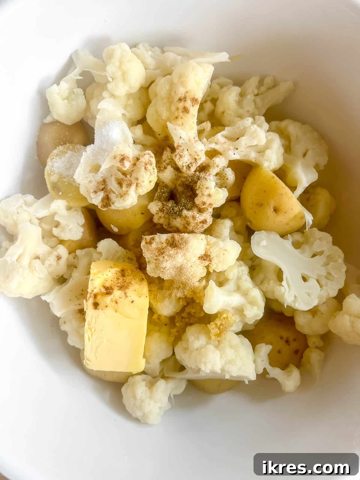 A large mixing bowl containing drained boiled potatoes and cauliflower, with all the additional ingredients like butter, half-and-half, and seasonings added on top, ready for mashing.