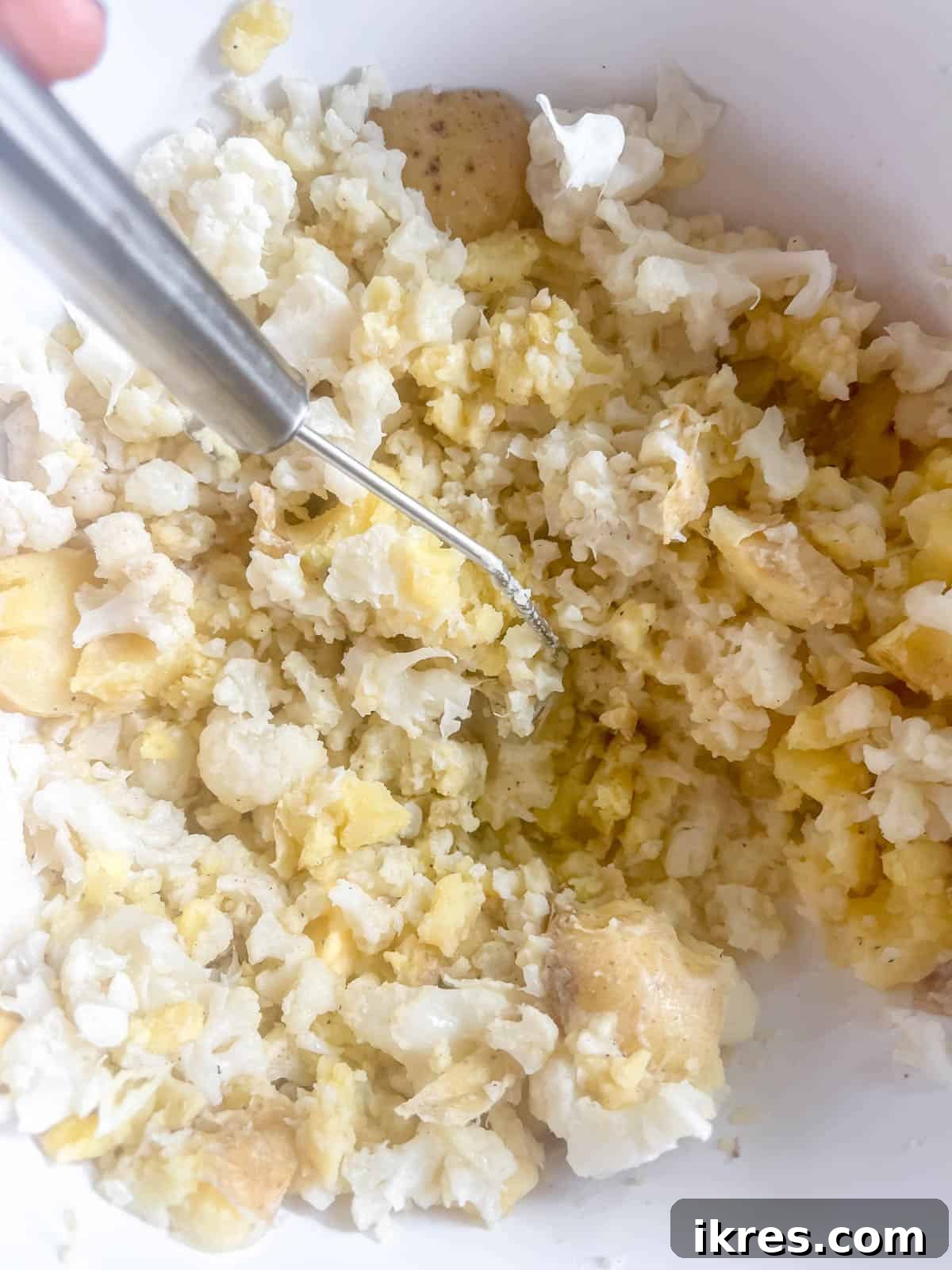 A potato masher actively mashing the combined cauliflower, potatoes, butter, half-and-half, and seasonings in a large bowl, creating a creamy texture.