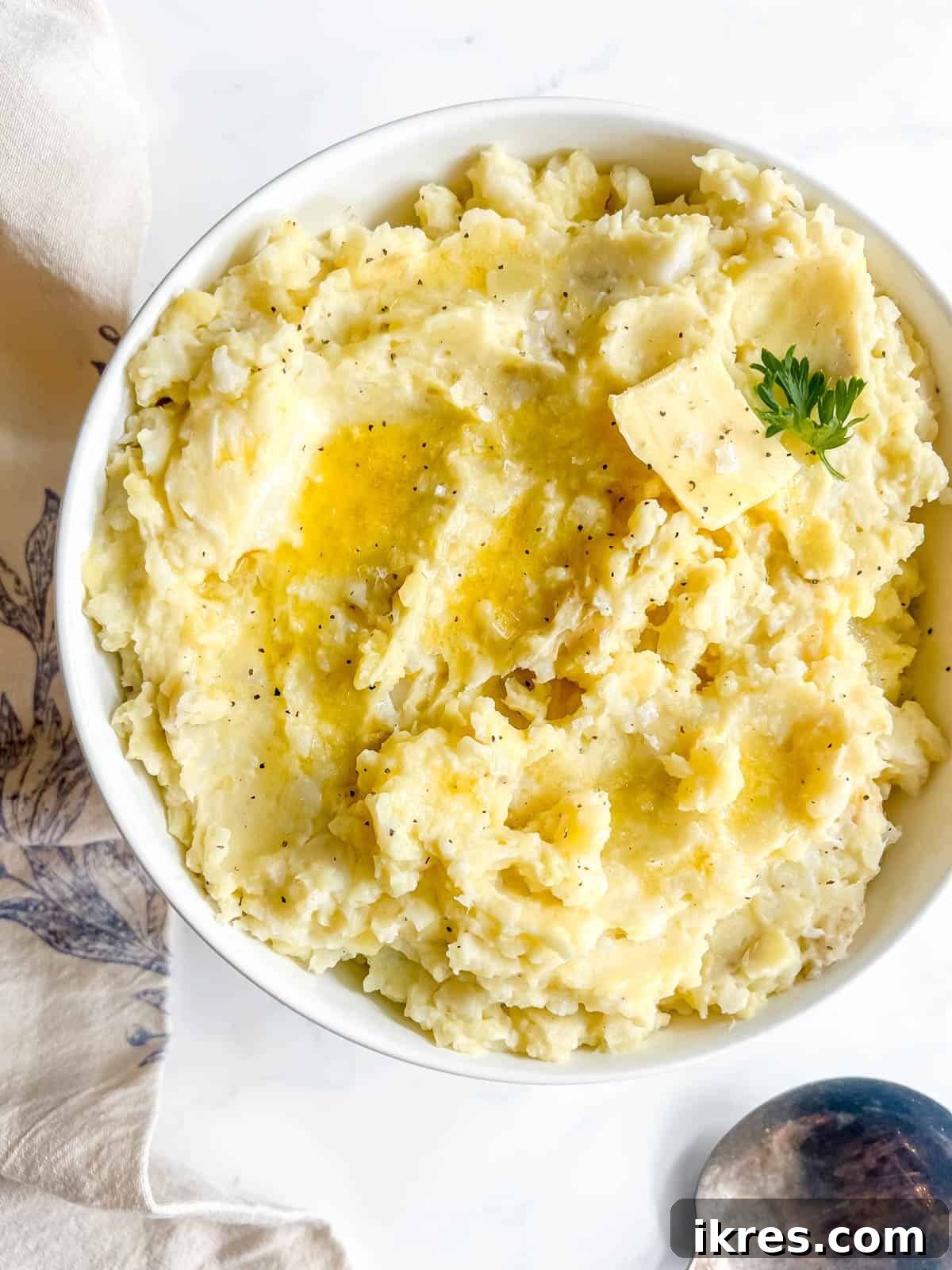 A serving bowl of finished cauliflower and potato mash, generously topped with a pat of melting butter and a sprinkle of fresh parsley, ready for the table.