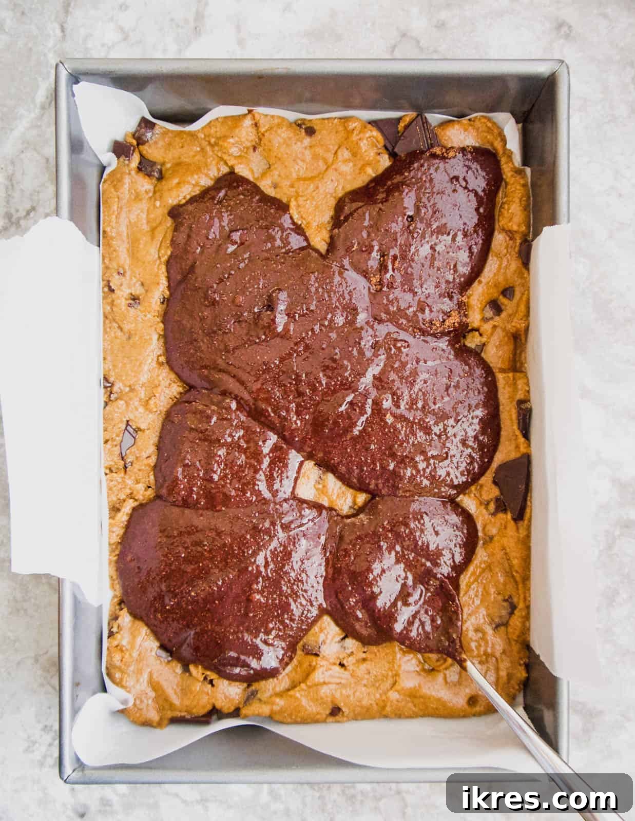 Thick brownie batter being carefully poured over the cookie layer in a baking pan.