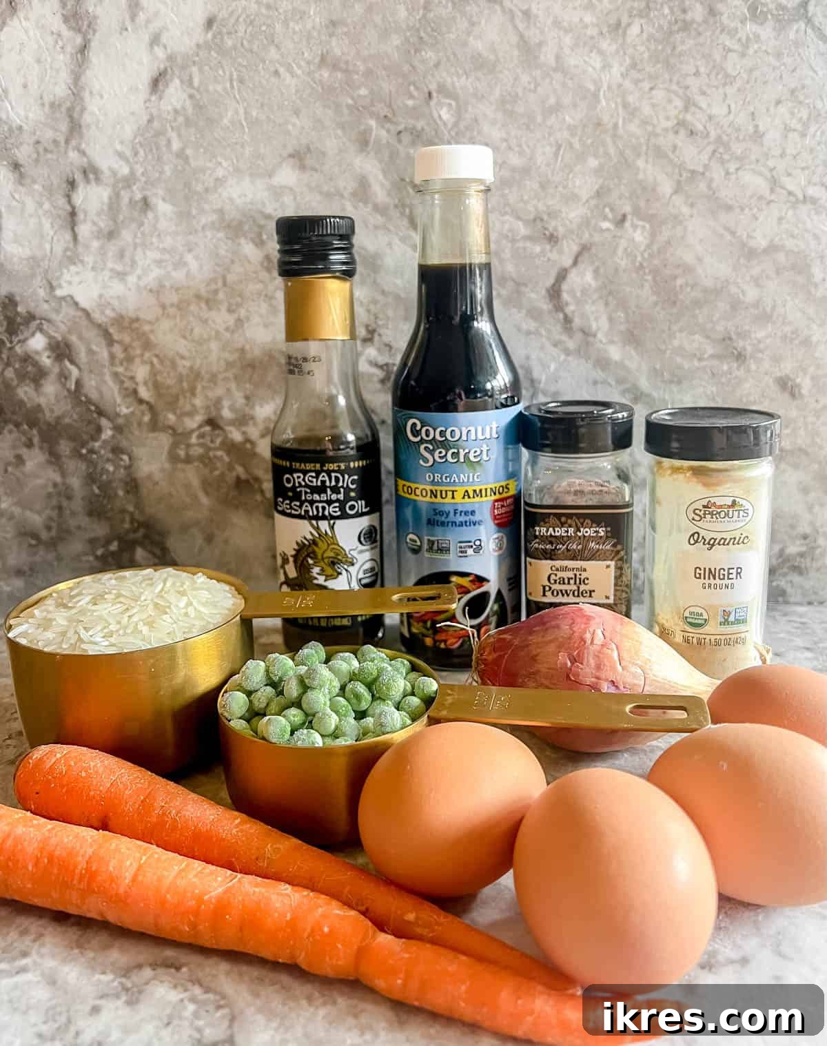 A flat lay photograph displays all the fresh ingredients meticulously arranged for making coconut aminos fried rice, including a bowl of cooked white rice, eggs, diced onion, peas, carrots, sesame oil, and a bottle of coconut aminos, alongside ginger and garlic powder.