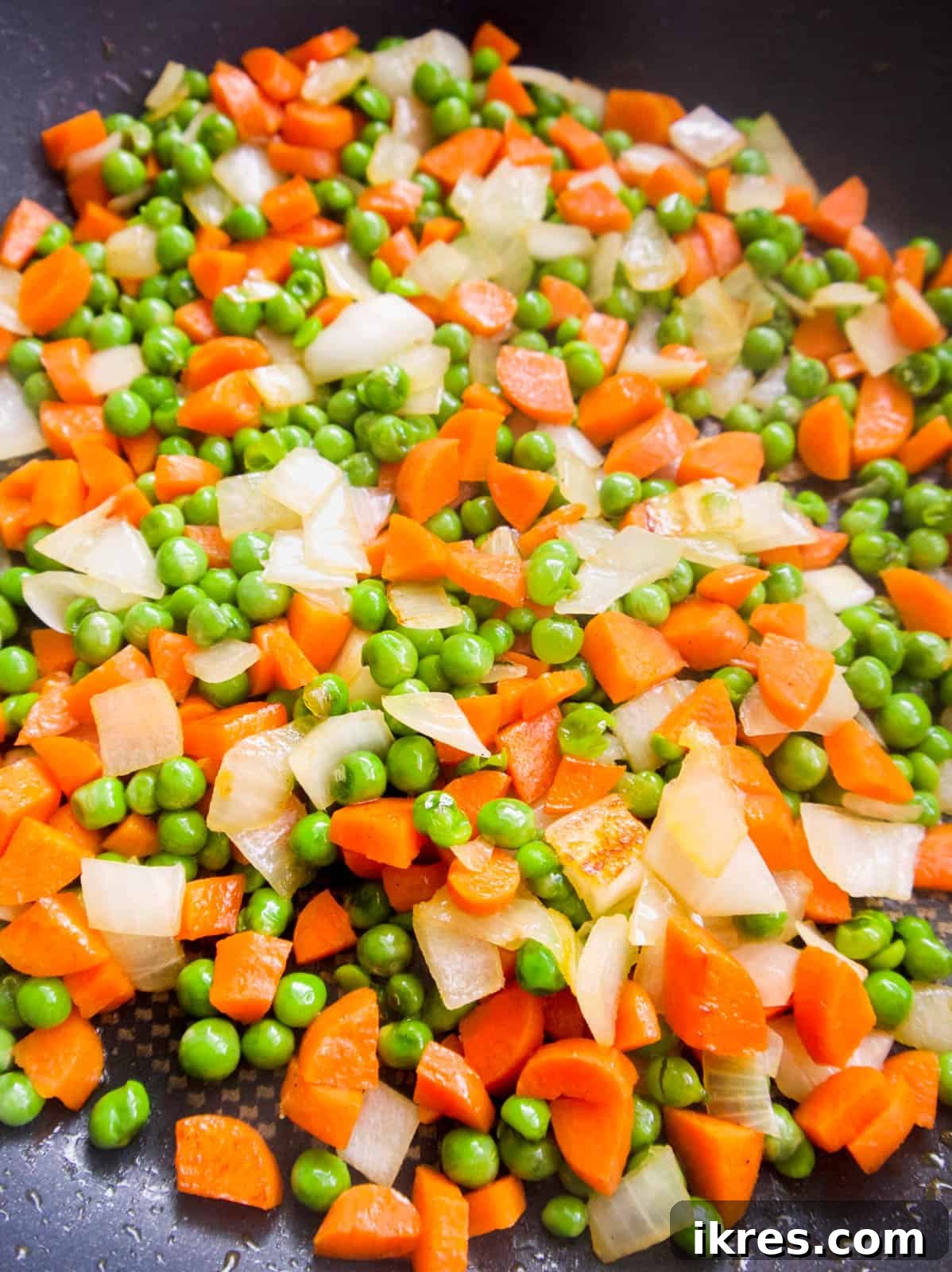 Diced white onion, vibrant green peas, and thinly sliced carrots are actively sautéing in glistening sesame oil within a large frying pan, emitting aromatic steam.