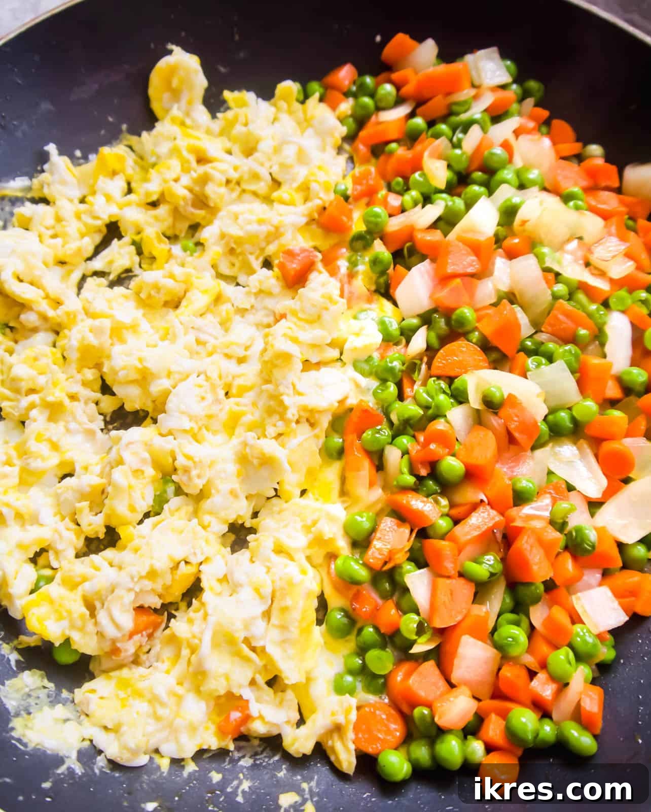 In a frying pan, half of the surface holds scrambled eggs cooking, while the other half contains the previously sautéed carrots and peas, separated and ready to be combined.