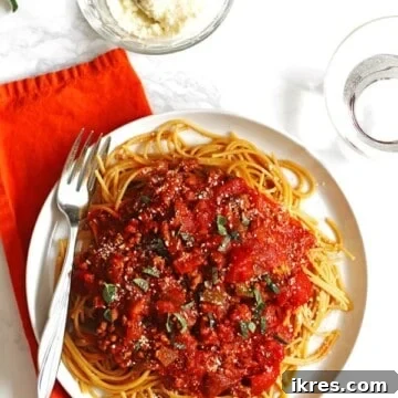 Farklı taze otlar ile süslenmiş beyaz bir tabakta spagetti sosu ve makarnalar. Yanında rendelenmiş parmesan peyniri kasesi ve bir kaşık bulunuyor.