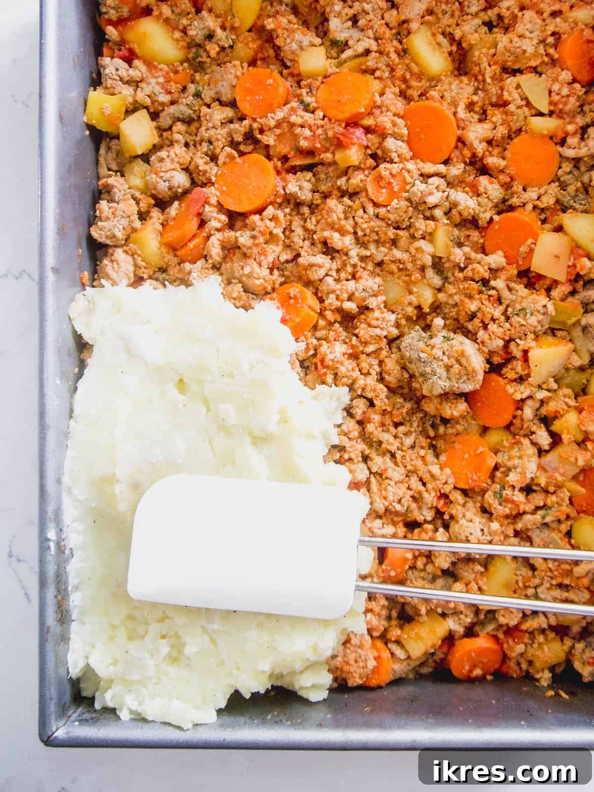Creamy mashed potatoes being spooned over the meat filling in the baking dish.