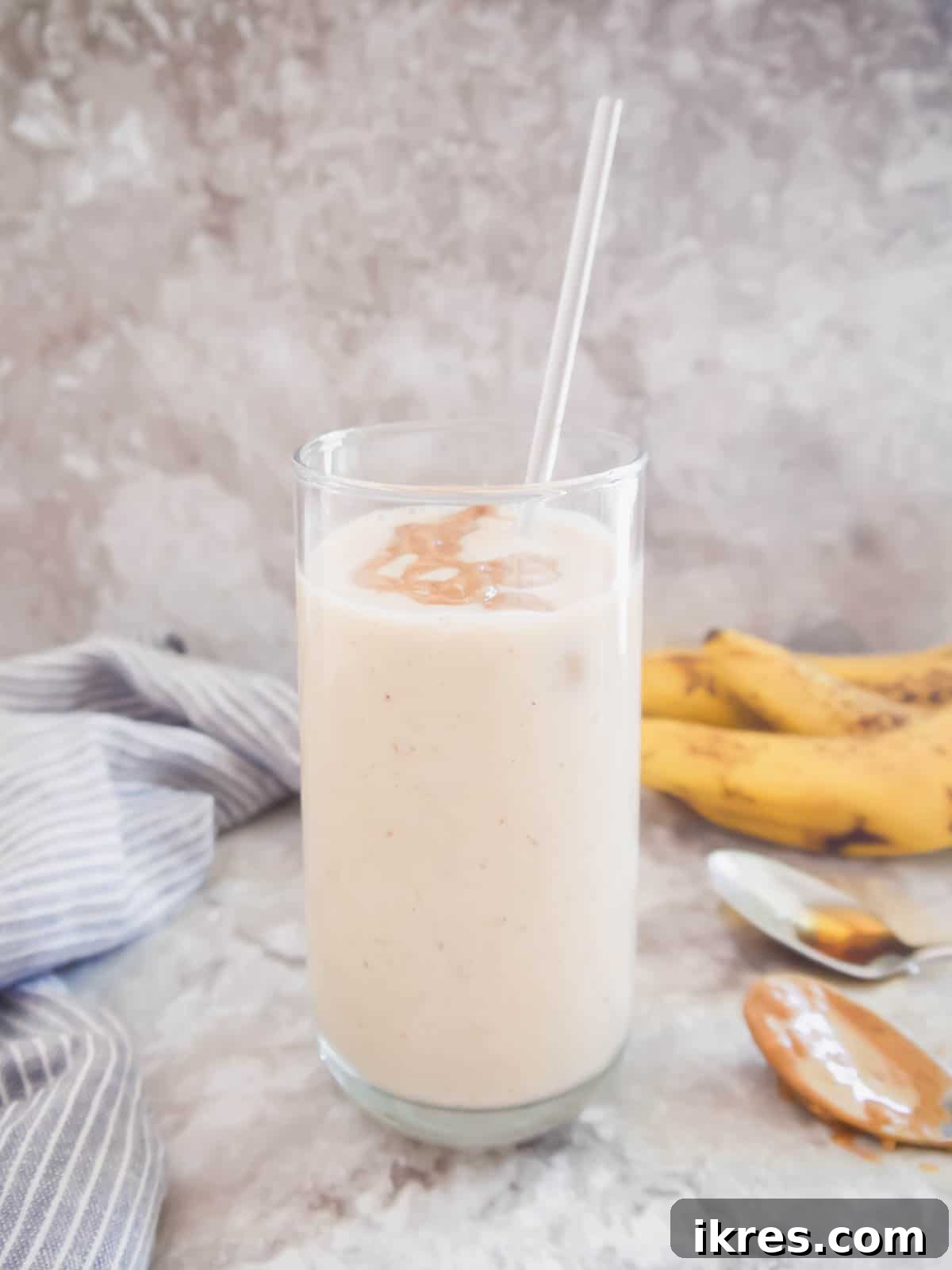 A tall glass of creamy banana yogurt smoothie with maple syrup, garnished with a straw and a fresh slice of banana, against a light background.