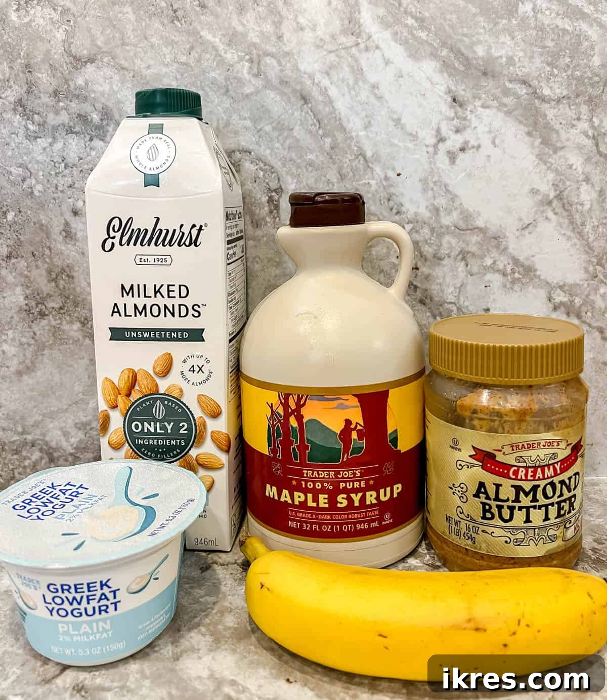A vibrant selection of smoothie ingredients including neatly sliced frozen bananas, a bottle of pure maple syrup, a tub of creamy yogurt, a carton of almond milk, and a jar of smooth almond butter, all arranged on a light surface.