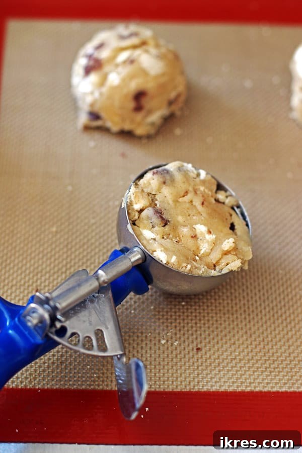 Toasted Smores Cookies 6 A large cookie scoop filled with S'mores cookie dough, showcasing its generous size, is poised above a baking sheet lined with parchment paper, ready to be placed for baking. This illustrates the precise portioning for jumbo cookies.