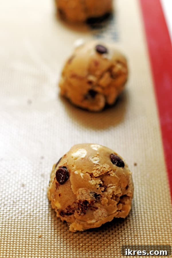 Toasted Smores Cookies 7 Six generously sized S'mores cookie dough balls are neatly arranged on a baking sheet lined with parchment paper, each topped with visible chocolate, marshmallow, and graham cracker pieces, ready for baking.