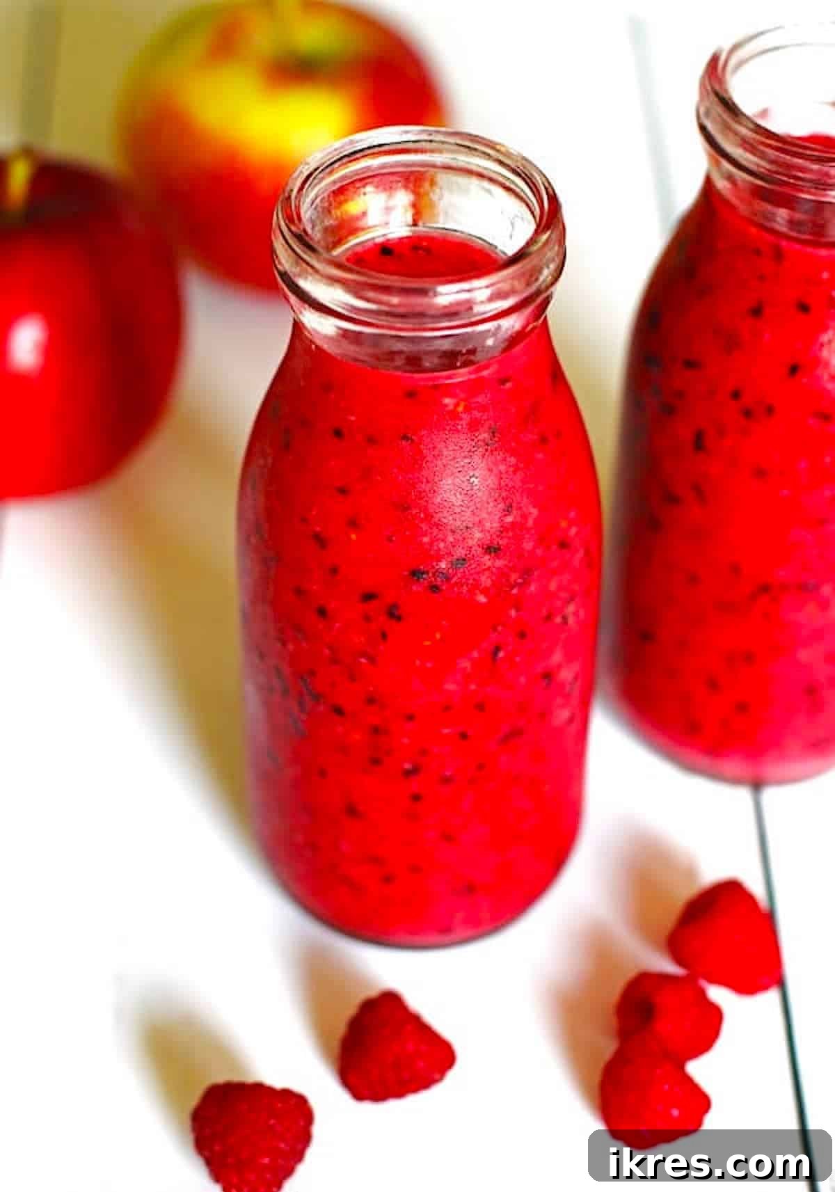 Berry Orchard Smoothie 2 Two clear glasses of Apple Smoothie on a white table, surrounded by fresh apples and raspberries.