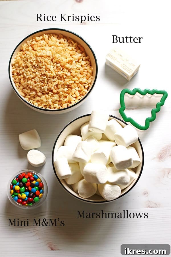 A selection of ingredients for Christmas Rice Krispie Treats, including a bowl of Rice Krispies cereal, a bowl of marshmallows, a small dish of mini M&M candies, and a stick of butter, arranged on a surface with a tree-shaped cookie cutter.