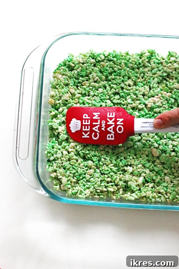 A hand using a buttered spatula to evenly spread a green Rice Krispie Treat mixture into a 13x9 inch baking pan.