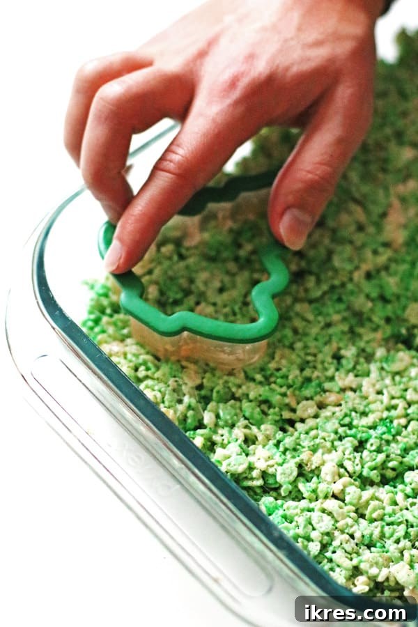 A tree-shaped cookie cutter is pressed into a pan of green Rice Krispie Treats to cut out festive shapes for Christmas.