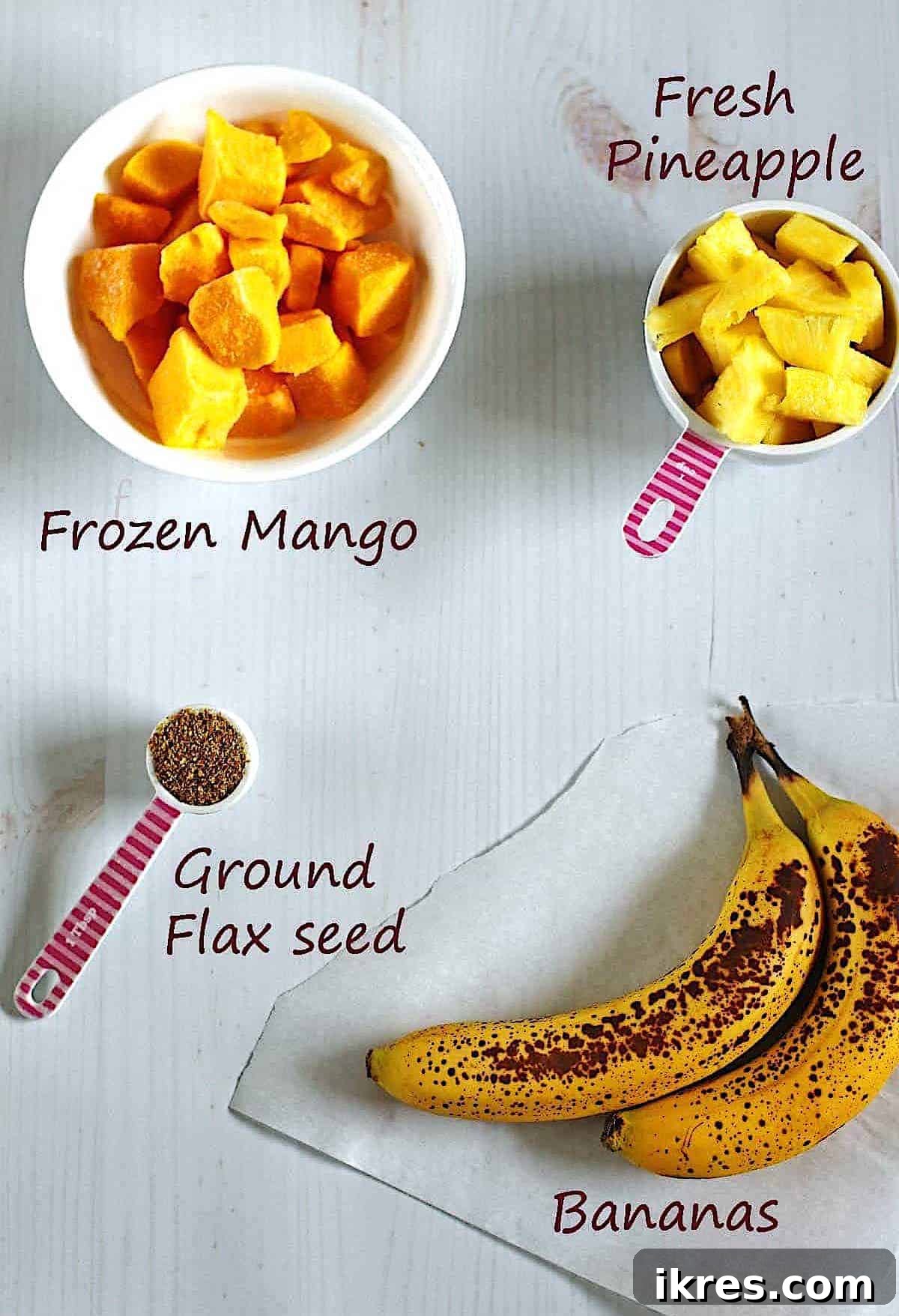 A vibrant selection of ingredients for a Pineapple Mango Smoothie: a bowl of frozen mango, fresh pineapple chunks, ground flaxseed, and two ripe bananas, all arranged on a clean white surface.