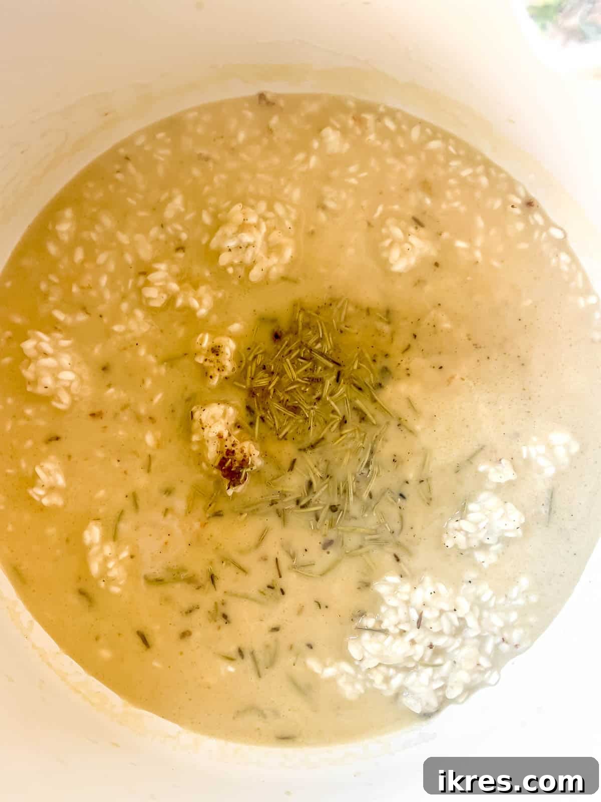 A mix of chicken broth and various spices like nutmeg and rosemary added to the risotto in a pan.
