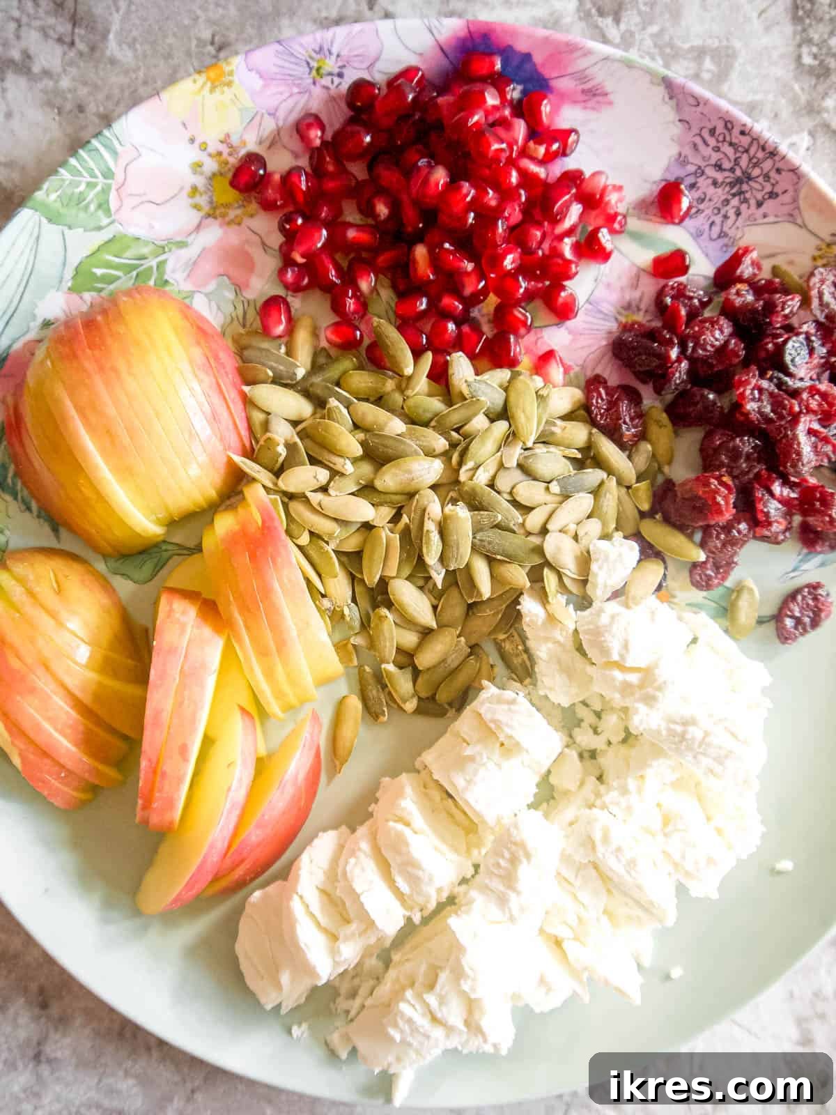 Various prepped salad ingredients including sliced apples, crumbled goat cheese, and pomegranate seeds neatly arranged on a plate.