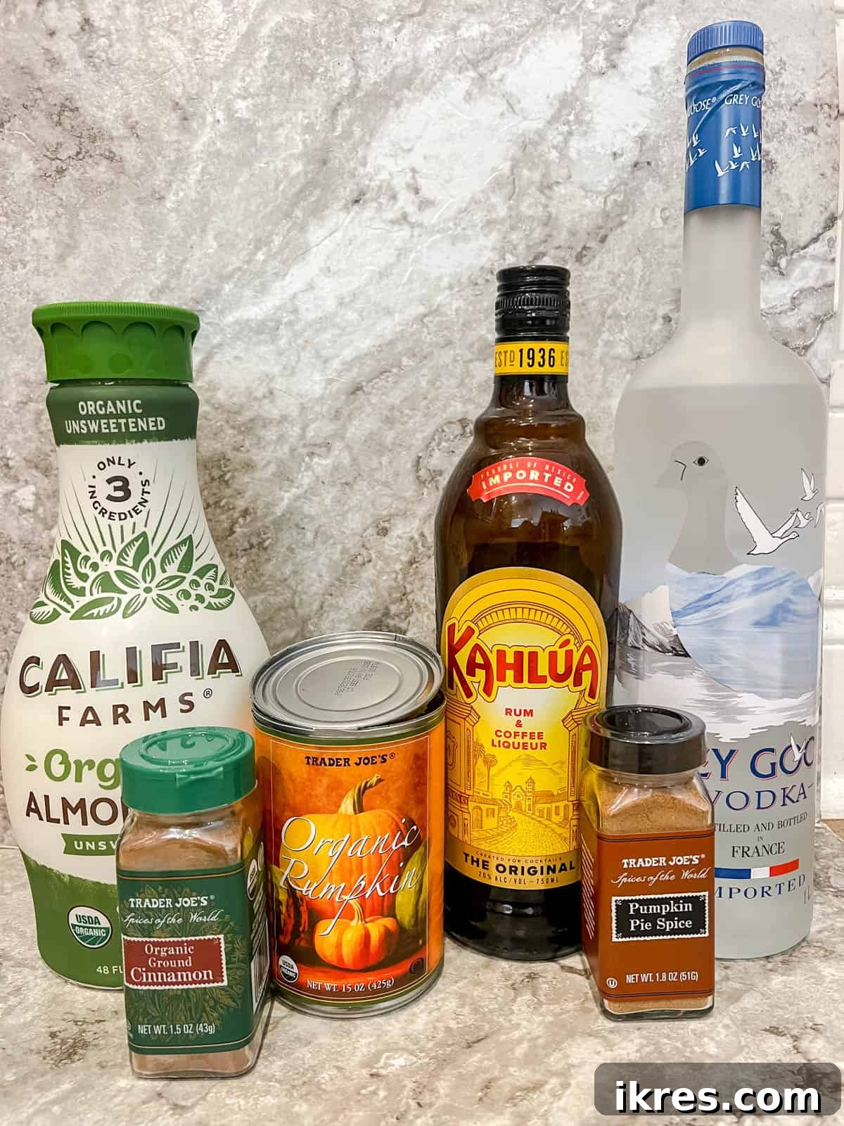 Ingredients for Pumpkin Spice White Russian arranged on a countertop, including almond milk, vodka, coffee liqueur, pumpkin puree, pumpkin pie spice, and maple syrup.